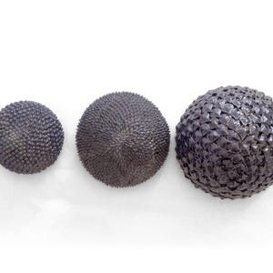 Puede incluir: Tres objetos decorativos esféricos de color gris oscuro de varios tamaños están dispuestos sobre un fondo blanco. Los objetos tienen superficies texturizadas, pareciendo el más grande tejido. La esfera más pequeña tiene una textura puntiaguda.