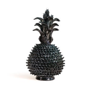 Puede incluir: Una escultura de cerámica negra brillante con forma de piña. El cuerpo presenta un patrón de escamas texturizado y repetitivo, mientras que la parte superior consta de hojas puntiagudas y curvas que irradian sobre una base circular.