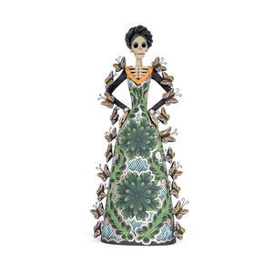 May include: A Day of the Dead figurine of a skeleton woman in a green floral dress adorned with butterflies. The figure has a black hairstyle and is wearing a black top with an orange collar. The dress features a detailed floral pattern.
