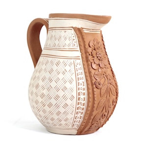 May include: A handcrafted ceramic pitcher with a cream-colored body and a brown handle and accents. The pitcher features a geometric pattern on the front and floral relief on the side, showcasing a rustic aesthetic.