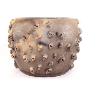 Op de afbeelding: Een ronde, grijsbruine keramische plantenbak met een getextureerd oppervlak. De plantenbak is versierd met talrijke kleine, verhoogde schedelontwerpen. De schedels zijn in verschillende tinten gebroken wit en grijs.