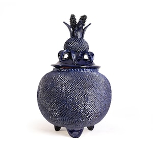 May include: A deep blue ceramic jar with a textured, cross-hatched surface pattern. The jar features a removable lid topped with a sculptural, pineapple-like finial and rests on three small, dark, rounded feet, creating a distinct silhouette.