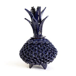 Puede incluir: Un tarro de cerámica azul oscuro con forma de piña y tapa extraíble. El cuerpo presenta un patrón texturizado de escamas con pequeños detalles florales en la parte superior. El recipiente se apoya sobre tres patas cortas y puntiagudas con un acabado brillante.