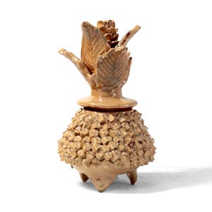 May include: A beige ceramic container with a distinctive design. It has a textured, rounded base, a decorative midsection, and a top with leaf-like elements. The piece is raised on three small feet. Measurements are in centimetres.
