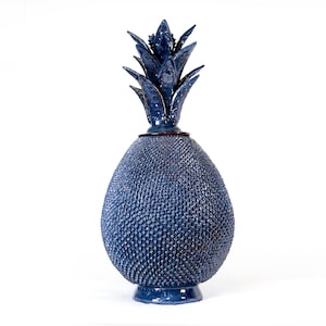Puede incluir: Un objeto decorativo de cerámica con forma de piña y un esmalte azul oscuro brillante. El cuerpo presenta una superficie texturizada con pequeños relieves y la parte superior consta de hojas puntiagudas erguidas sobre una base circular.
