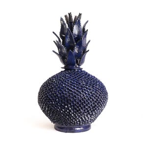 Puede incluir: Una escultura de piña de cerámica azul oscuro que presenta una superficie muy texturizada en forma de escamas en el cuerpo redondeado y una corona de hojas puntiagudas y dentadas en la parte superior. La pieza tiene un acabado brillante y reflectante.