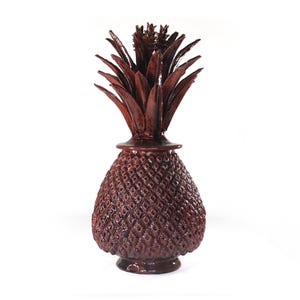 May include: A decorative, reddish-brown pineapple-shaped ornament. The pineapple has a textured surface and a detailed crown of leaves. The base is round and the object is set against a white background.