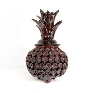 May include: A dark brown decorative pineapple-shaped container with a lid. The container is covered in small, flower-like details. The top has a leafy crown, and the base has three small feet. The background is white.