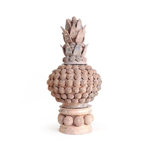 May include: A light brown terracotta decorative finial featuring a pineapple-shaped body covered in small, circular, textured rosettes. The top is adorned with pointed, leaf-like petals, and the base is a cylindrical pedestal with rosettes.