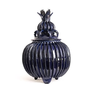 May include: A deep navy blue ceramic lidded jar with a glossy finish. The body features vertical ribbed detailing and a rounded shape. The lid is topped with a decorative, pointed, petal-like finial. The jar stands on three small, short feet.