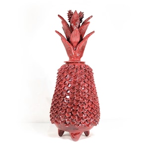 May include: A red ceramic pineapple-shaped decorative object. The pineapple has a textured, scale-like pattern on the body and a detailed leafy crown. It stands on three small feet and is a vibrant accent piece.