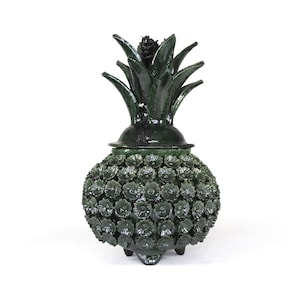 Puede incluir: Un recipiente decorativo en forma de piña de color verde oscuro con tapa y tres patas pequeñas. El cuerpo está cubierto de pequeños detalles en forma de flor. La parte superior presenta un grupo de hojas verdes.