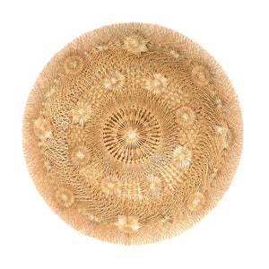 May include: A circular, woven decorative piece in a natural beige hue. The intricate design features concentric circles and star-like patterns, creating a textured surface. The craftwork is set against a white background.