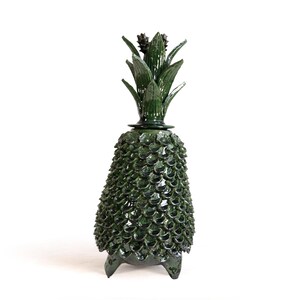 May include: A dark green ceramic pineapple-shaped jar with a removable lid. The body features a textured, overlapping scale pattern, while the lid is topped with sculpted, pointed leaves. The entire piece has a glossy finish and stands on three small feet.