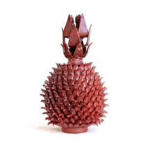 May include: A glossy, dark reddish-brown ceramic sculpture shaped like a pineapple. The body is covered in numerous pointed, overlapping leaf-like textures, and the top features a crown of upright, sharp, pointed ceramic leaves.