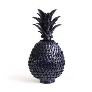 May include: A dark navy blue ceramic pineapple sculpture with a glossy finish. The object features a textured, scale-like pattern across the main body and a pointed, leafy crown at the top, standing on a small, circular base.