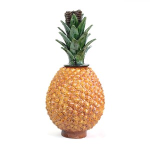 Könnte beinhalten: Dekoratives Objekt in Form einer Ananas aus Keramik. Der Körper ist strukturiert, goldbraun, mit einem grünen, blattartigen Oberteil und einer dunkelbraunen Krone. Der Sockel ist dunkler braun. Auf weißem Hintergrund.