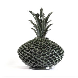 Puede incluir: Un objeto decorativo en forma de piña de color verde oscuro. El cuerpo tiene una textura escamosa y la parte superior presenta detalles de hojas. La piña está hecha de un material oscuro y metálico y se encuentra sobre un fondo blanco.