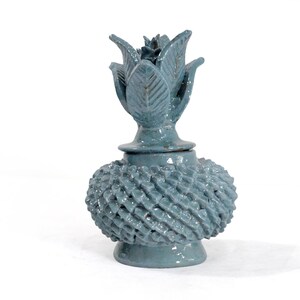May include: A blue-green ceramic decorative jar with a textured, woven-like spherical body and a floral-shaped lid. The jar has a pedestal base and a matching floral top. The overall height is approximately 7 inches.