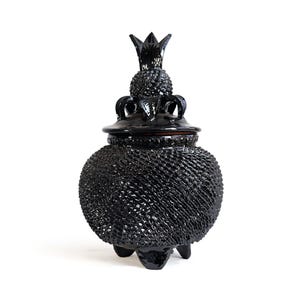 May include: A glossy black ceramic lidded jar with a textured, pineapple-like surface pattern. The lid features a decorative crown-shaped finial. The jar is supported by three small, sturdy feet at the base, creating a distinct silhouette.