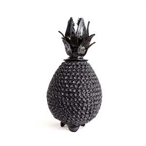 May include: A black decorative pineapple-shaped object with a textured, repeating circular pattern on the main body and a pointed, leaf-like top section. The object stands on small, integrated feet and features a glossy, uniform black finish.