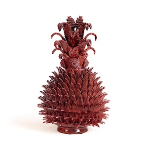 Peut inclure: Une sculpture en céramique rouge foncé en forme d'ananas, présentant une surface très texturée avec de nombreuses écailles pointues et superposées couvrant le corps et une couronne de feuillage détaillé et dressé sur une base.