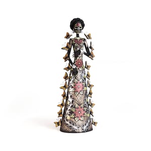May include: A tall, slender figurine of a skeletal figure with a dark complexion, wearing a long dress adorned with pink floral patterns and silver filigree. Numerous small, golden-brown butterflies are attached to the sides of the figure.