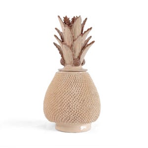 May include: A decorative pineapple-shaped ceramic container. The container is light beige with a textured, dotted surface. The top features a detailed, spiky crown in shades of brown and beige. The base is a lighter beige colour.