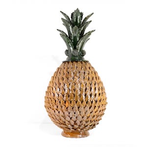 Puede incluir: Una gran escultura decorativa de piña. El cuerpo es de color marrón dorado texturizado, con un patrón de formas superpuestas en forma de hoja. La parte superior presenta hojas de color verde oscuro y una pequeña base verde oscuro.