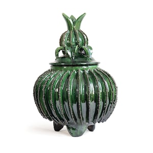 May include: A dark green ceramic lidded jar with a glossy finish. The body features vertical ribbed textures with small bead-like details along the ridges. It stands on three short feet and includes a matching lid topped with a pointed finial.