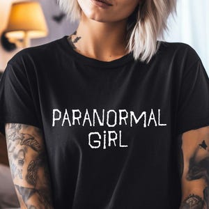 May include: Black t-shirt with the text "PARANORMAL GIRL" in white. The shirt is a crew neck style. The background features a wall decorated with bat silhouettes.