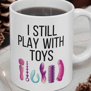 May include: White ceramic coffee mug with a black handle and the text "I still play with toys" printed in black. The mug has a row of colorful toy illustrations printed below the text.