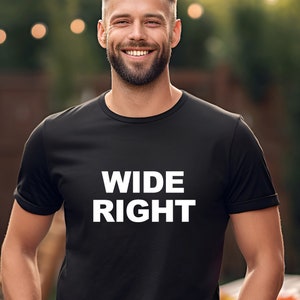 May include: Black crew neck t-shirt with the words "WIDE RIGHT" in bold white letters. The shirt is worn by a person standing outdoors with a blurred background of a wooden fence, trees, and a table with fruit.