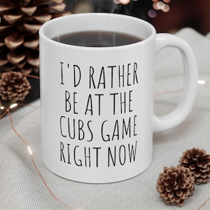 May include: White ceramic mug with the text "I'D RATHER BE AT THE CUBS GAME RIGHT NOW" in black. The mug is filled with a dark liquid and has a curved handle. The mug is on a light gray surface with decorative elements.