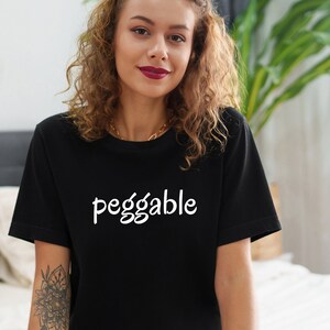 Può includere: Una maglietta nera con la scritta "peg gable" stampata in bianco sul davanti. La persona che indossa la maglietta è seduta su un letto con le gambe incrociate.