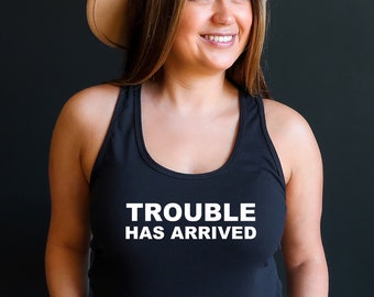 May include: Black racerback tank top with the white text "TROUBLE HAS ARRIVED". The person in the image is wearing a tan hat and light blue denim shorts. The background is a solid black.