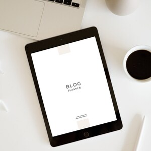 May include: A black tablet displaying a white screen with the words "BLOG PLANNER" in a minimalist font. The tablet is on a white surface, next to a laptop, a cup of coffee, and a pen. The text "STOP DREAMING AND START DOING" is also visible.