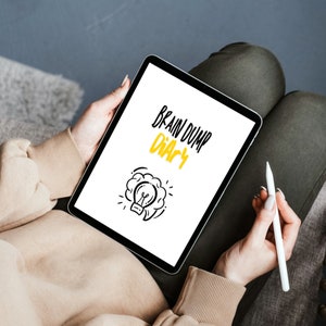 Könnte beinhalten: Ein weißes Tablet-Computer mit schwarzem Rand, auf dem der Text "Brain Dump Diary" mit einer Glühbirnengrafik angezeigt wird. Eine Person hält das Tablet und einen weißen Stift.