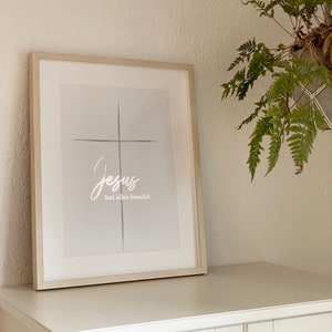 Könnte beinhalten: Ein gerahmter Druck mit einem weißen Kreuz und dem Text "Jesus hat alles bezahlt" in weißer Kursivschrift auf hellgrauem Hintergrund.