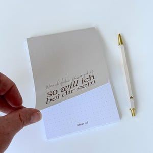 Könnte beinhalten: Ein Notizblock mit dem Text "Wenn du durchs Wasser gehst, SO will ich bei dir sein" und "Hebräer 13,5" aufgedruckt. Ein weißer Stift mit goldenen Akzenten und einem Herzdetail liegt neben dem Notizblock.