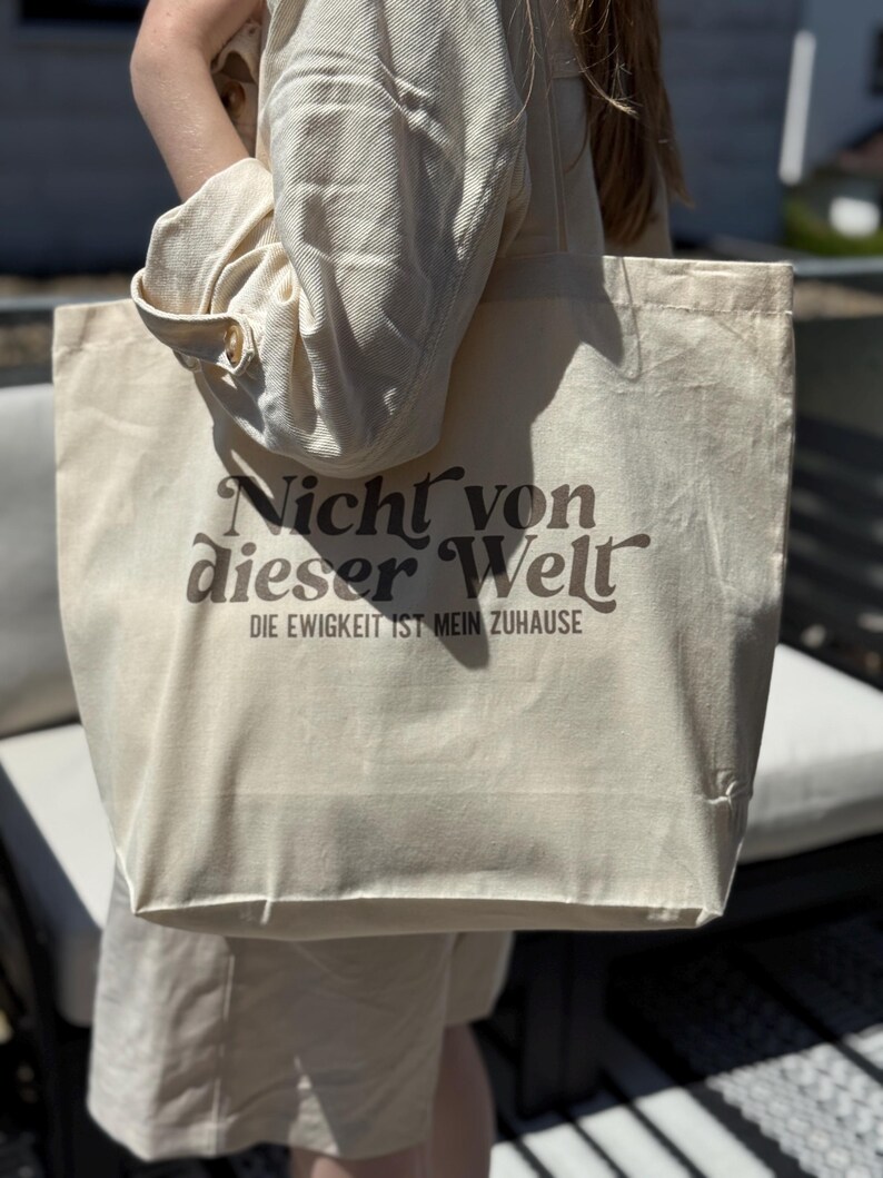 Może przedstawiać: Kremowa torba typu tote z brązowym napisem "Nicht von dieser Welt" i "DIE EWIGKEIT IST MEIN ZUHAUSE". Torba wykonana jest z wytrzymałej tkaniny i ma prosty, minimalistyczny design. Torba jest trzymana przez osobę ubraną w kremowy str&oacute;j.