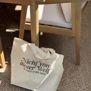 Może przedstawiać: Kremowa torba typu tote z napisem "Nicht von dieser Welt" i "Die Ewigkeit ist mein Zuhause" wydrukowanym w kolorze ciemnobrązowym. Torba ma dwa uchwyty i spoczywa na tkanej macie w pobliżu drewnianych mebli ogrodowych.