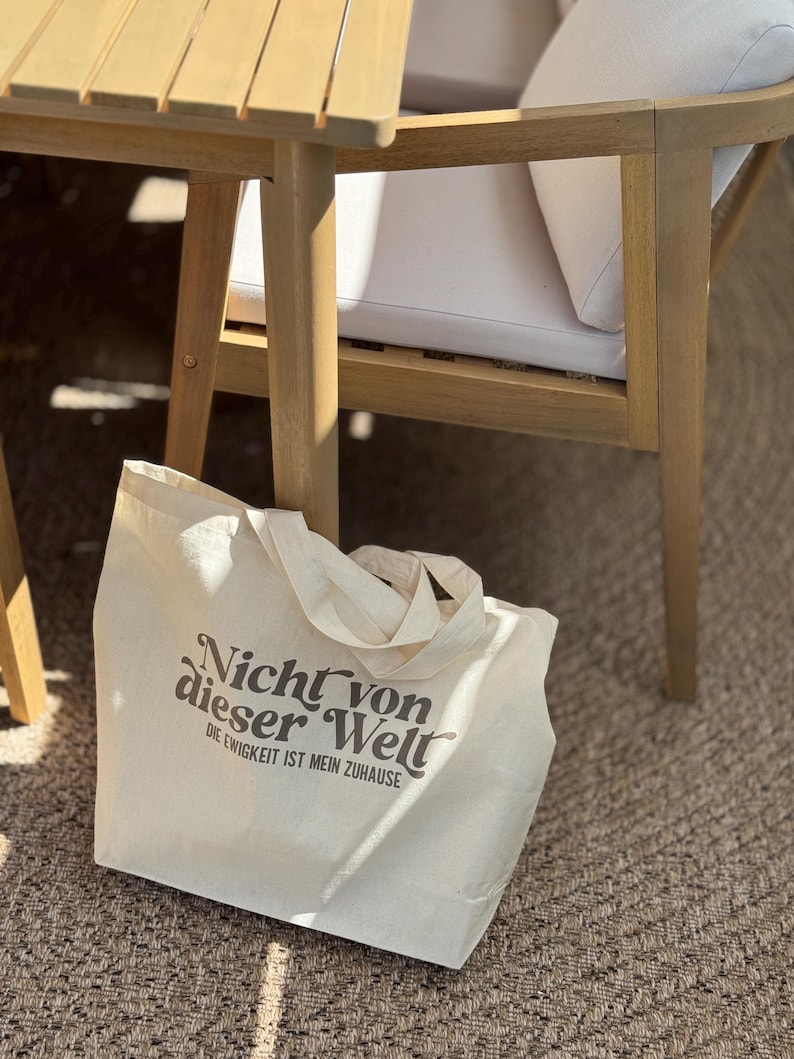 Może przedstawiać: Kremowa torba typu tote z napisem "Nicht von dieser Welt" i "Die Ewigkeit ist mein Zuhause" wydrukowanym w kolorze ciemnobrązowym. Torba ma dwa uchwyty i spoczywa na tkanej macie w pobliżu drewnianych mebli ogrodowych.