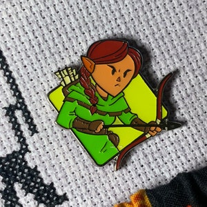 May include: An enamel pin depicting a cartoon elf with red hair, wearing a green hooded cloak and holding a bow and arrow.