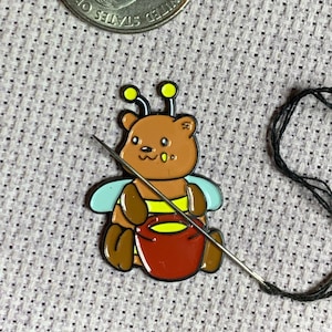 Puede incluir: Un oso marrón que lleva un disfraz de abeja con rayas amarillas y negras, alas azules y antenas amarillas. El oso sostiene una olla roja de miel. Una aguja e hilo negro están en el fondo.