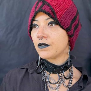 May include: A burgundy and black crochet beanie with a geometric design. The hat is paired with a black leather choker featuring large metal rings and chains. The overall aesthetic is dark and edgy.