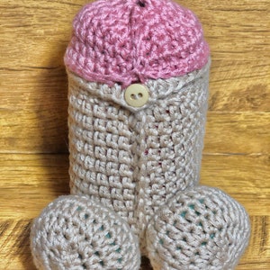 May include: A crocheted plush toy shaped like a human body. The toy is beige with a pink crocheted hat and a button on the front. The toy has two beige crocheted balls for feet.