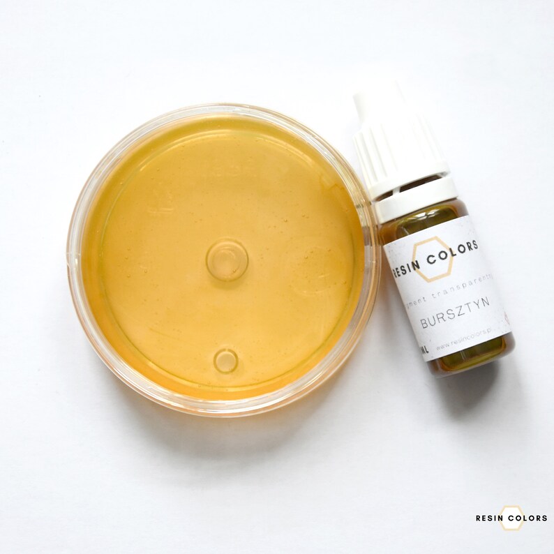 May include: A clear, round container filled with amber-colored liquid next to a small bottle. The bottle has a white cap and a label that reads "RESIN COLORS" and "BURSZTYN". The liquid in the container appears smooth and reflective.