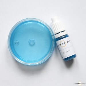 May include: A clear plastic container filled with light blue liquid next to a small bottle of blue resin. The bottle has a white cap and a label that reads "RESIN COLORS" and "BŁĘKIT".