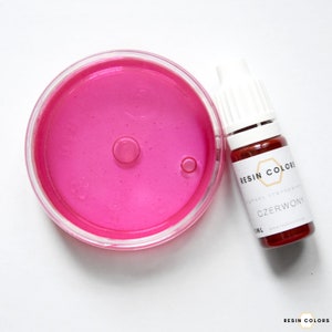 May include: A clear, round container filled with bright pink liquid next to a small bottle of red liquid. The bottle has a white cap and a label that reads "CZERWONY" and "RESIN COLORS".